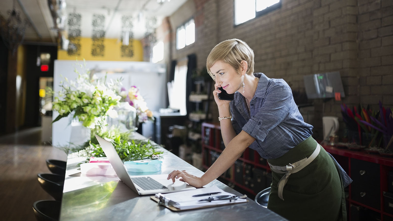 A woman in a shop, acknowledged as Google Play merchant, working on her laptop and talking to a client on the phone.