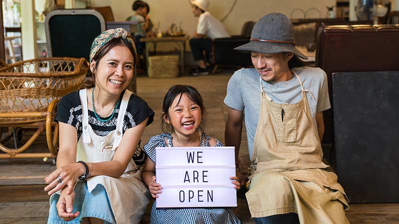 A family of three displays a "We are open" sign while sitting on the floor, representing the need of support for local businesses.